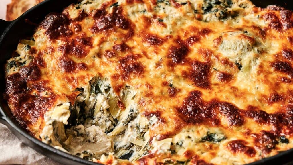 Close-up of a baked spinach artichoke dip in a black dish, with a portion scooped out showing the creamy interior.