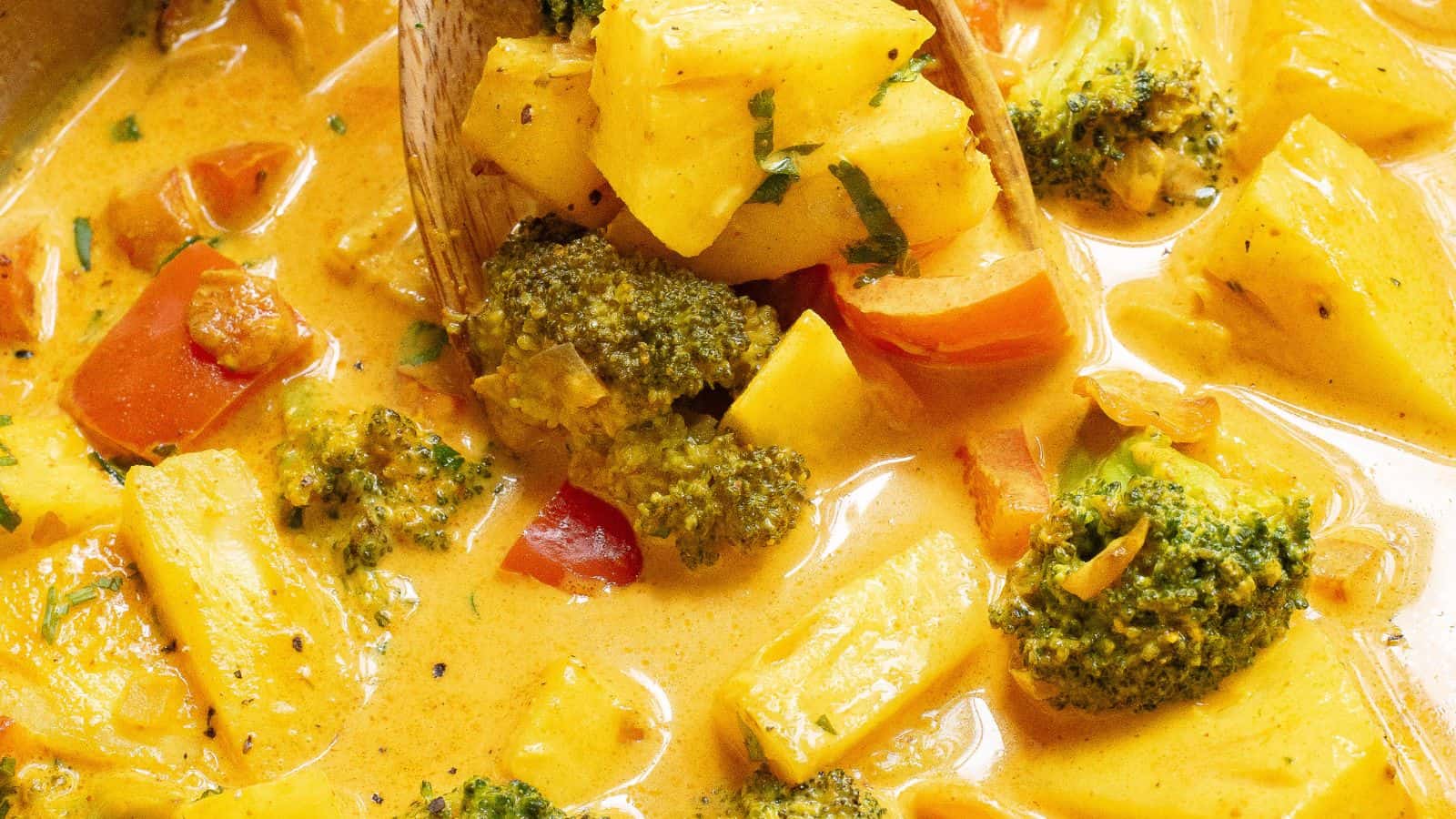 Close-up of a creamy yellow curry dish with broccoli, pineapple, and red bell peppers, stirred with a wooden spoon.