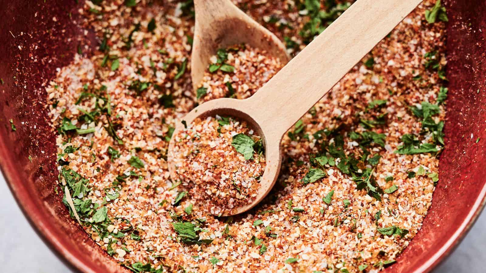 A wooden spoon rests in a bowl filled with a colorful spice mixture, including herbs like cilantro or parsley, with visible red and green flakes.