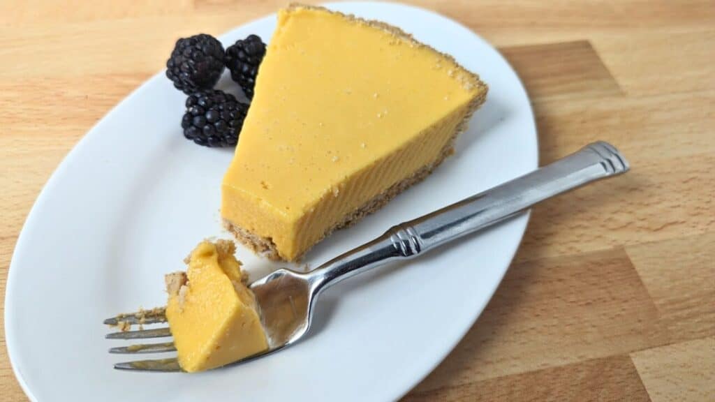 A slice of mango pie on a white plate with three blackberries and a fork holding a small piece of pie.