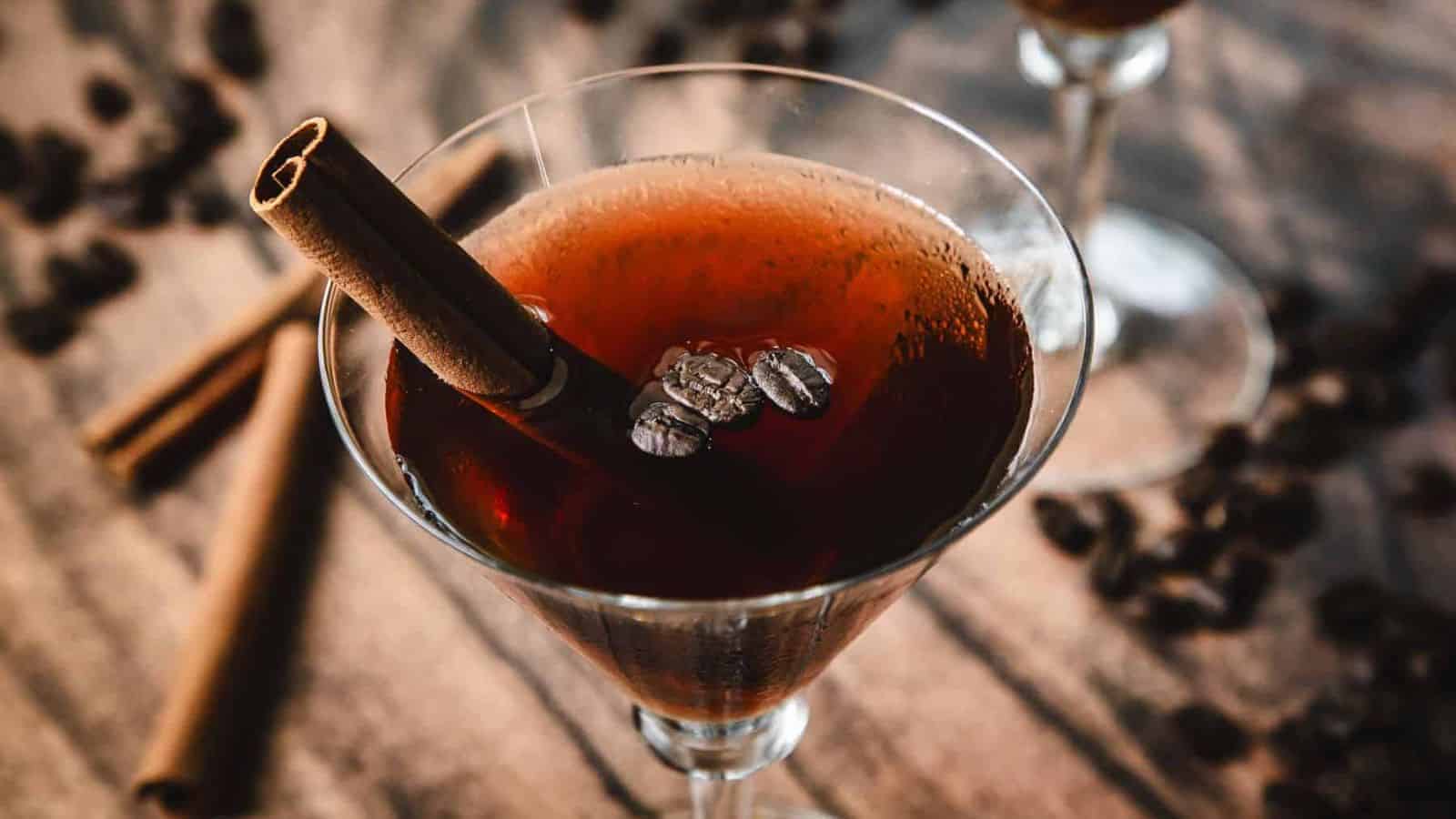 A martini glass filled with a dark brown cocktail, garnished with coffee beans and a cinnamon stick. Blurred cinnamon sticks and coffee beans are scattered on the wooden surface in the background.