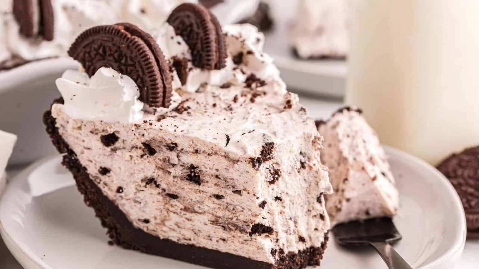 Slice of Oreo cream pie on a white plate, topped with whipped cream and Oreo cookies, with a fork resting beside it.