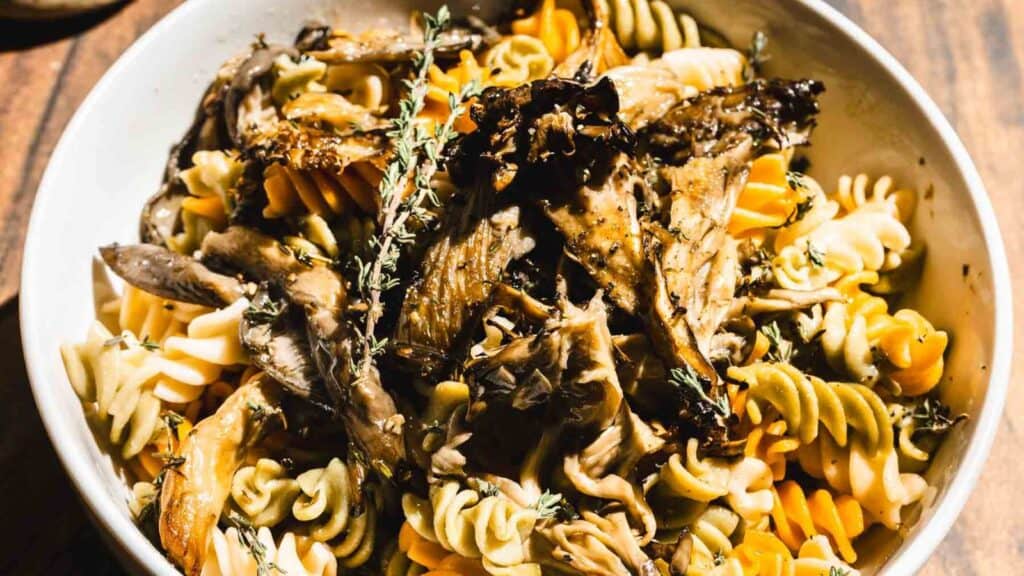 A bowl of tri-color fusilli pasta topped with roasted mushrooms and sprigs of thyme.