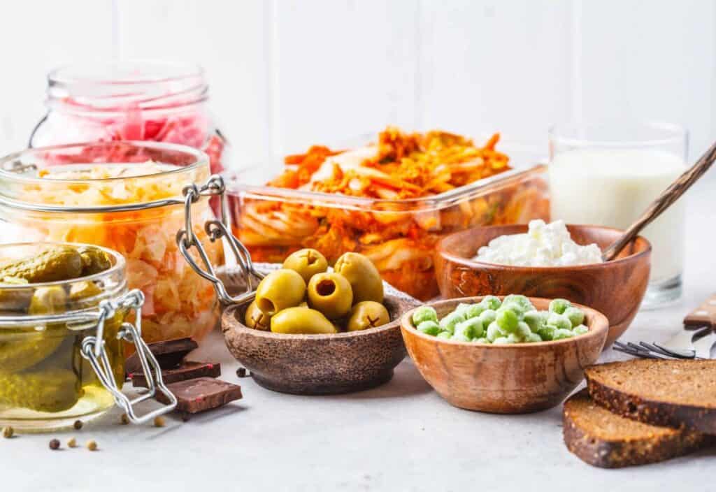 Assorted fermented foods, including sauerkraut, kimchi, pickles, olives, edamame, yogurt, and bread slices on a white surface&mdash;perfect for a winter blues cure.