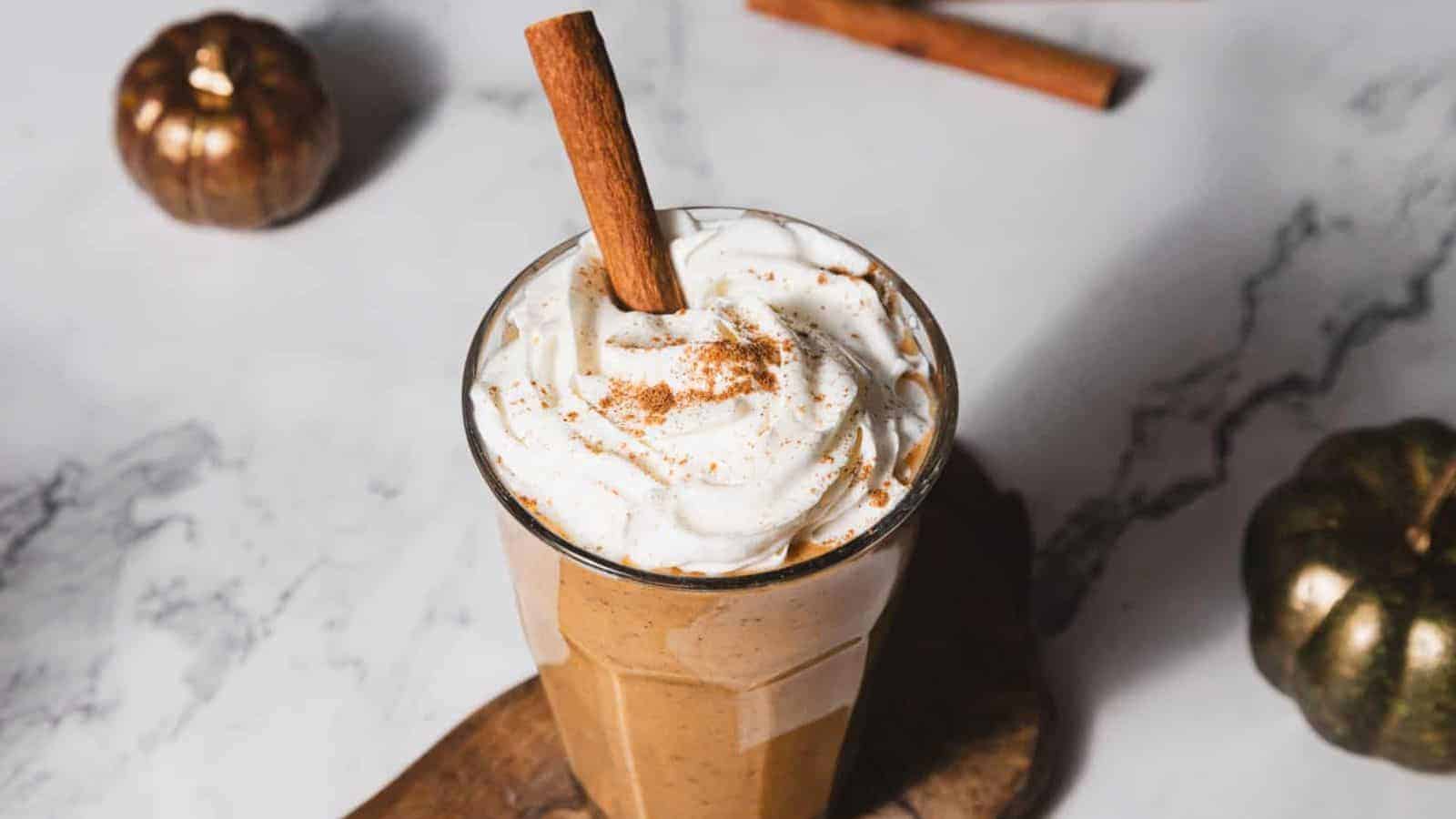 A tall glass filled with a creamy pumpkin spice latte topped with whipped cream and a cinnamon stick. The drink is set on a wooden coaster, and small decorative pumpkins are visible in the background on a marble surface.