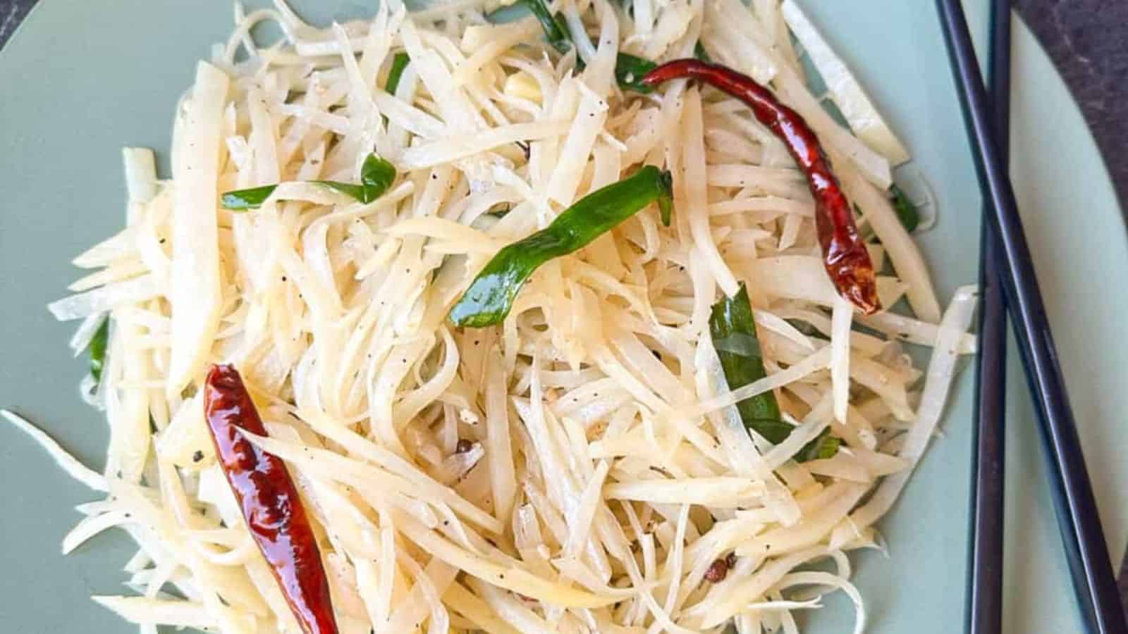 Stir-fried shredded potatoes with green onions and dried red chilies on a green plate, accompanied by black chopsticks.