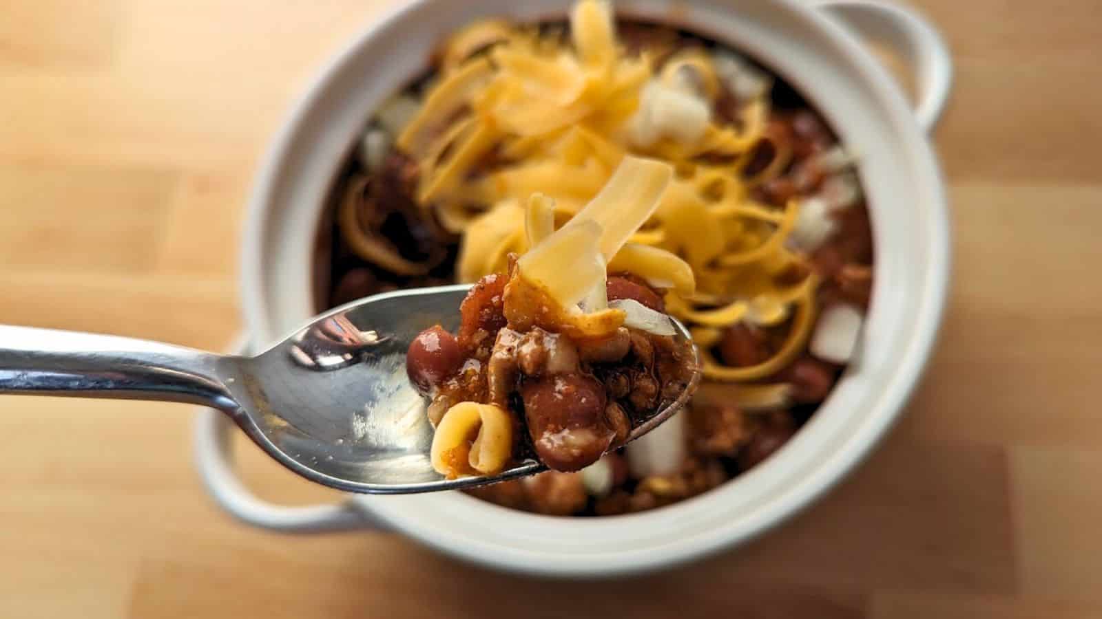 Image shows close-up of a spoonful of chili with beans, cheese, and corn, held above a pot filled with the same dish, placed on a wooden surface.