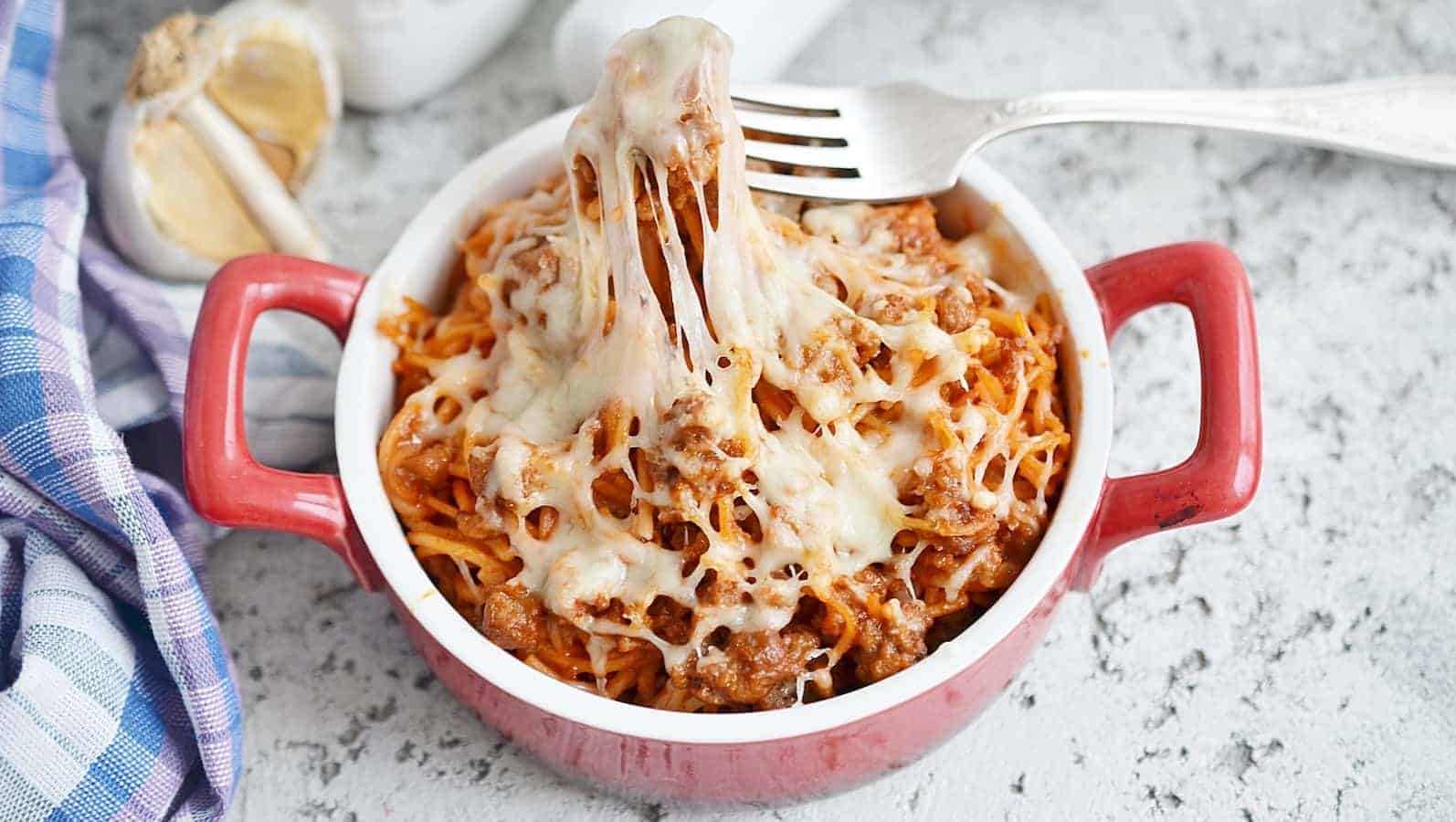 Baked spaghetti with melted cheese is served in a red dish, accompanied by a fork. Garlic and a checkered cloth are in the background.