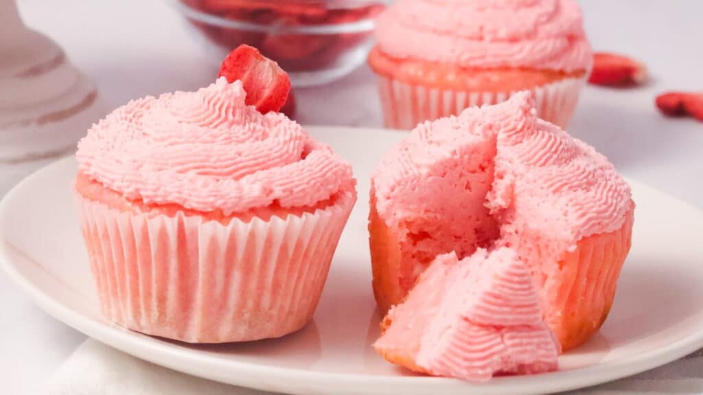 Two pink frosted strawberry cupcakes are on a white plate. One cupcake is intact, and the other has a piece cut out. A glass bowl with red ingredients is visible in the background.