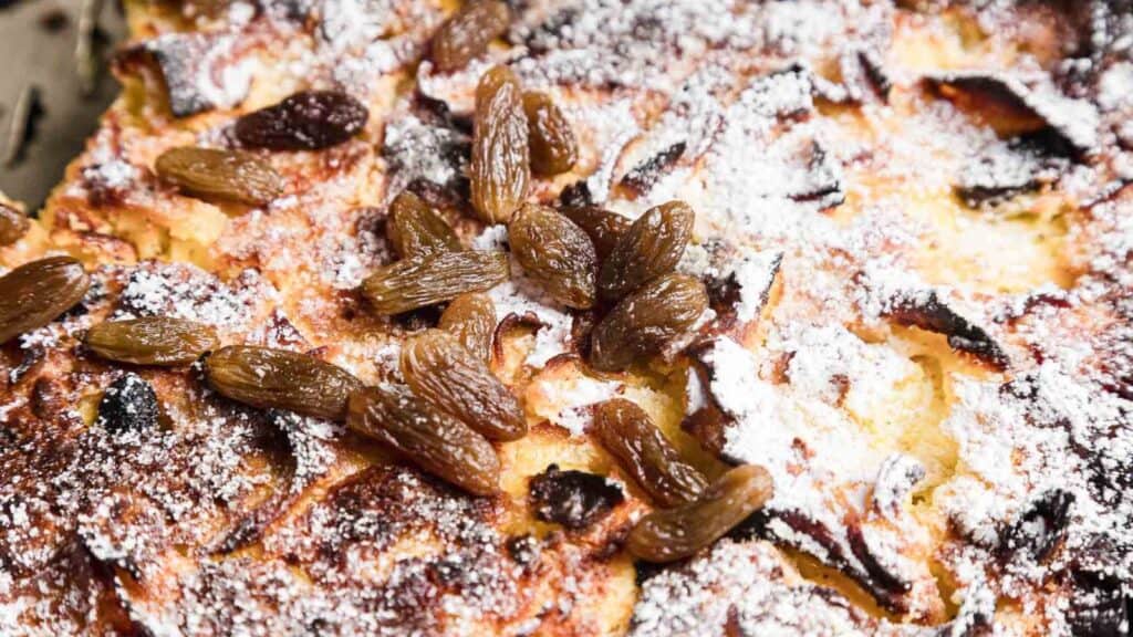 Close-up of a baked dessert topped with powdered sugar and raisins, featuring a crispy, browned surface.