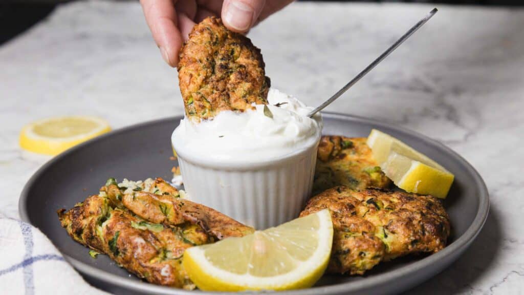 Person dips a fritter into a white sauce in a small ramekin on a plate with more fritters and lemon wedges.