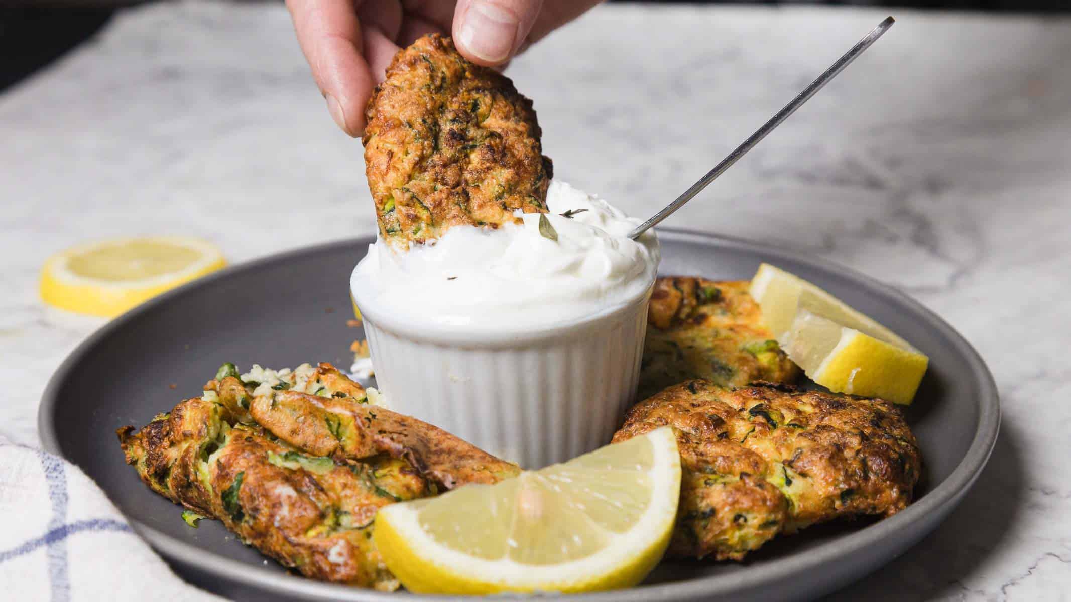 A hand dips a fried zucchini fritter into a small bowl of white cream sauce on a gray plate. Other fritters and lemon wedges are arranged around the bowl on a marble surface.