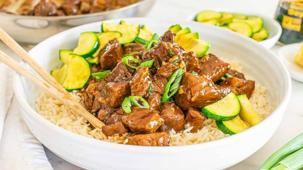 A bowl of rice topped with cubed steak bites in a brown sauce, garnished with sliced green onions and zucchini.