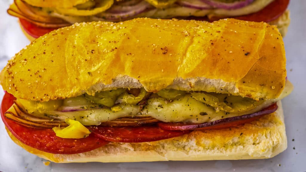Close-up of a toasted sandwich roll filled with salami, melted cheese, red onions, and banana peppers.