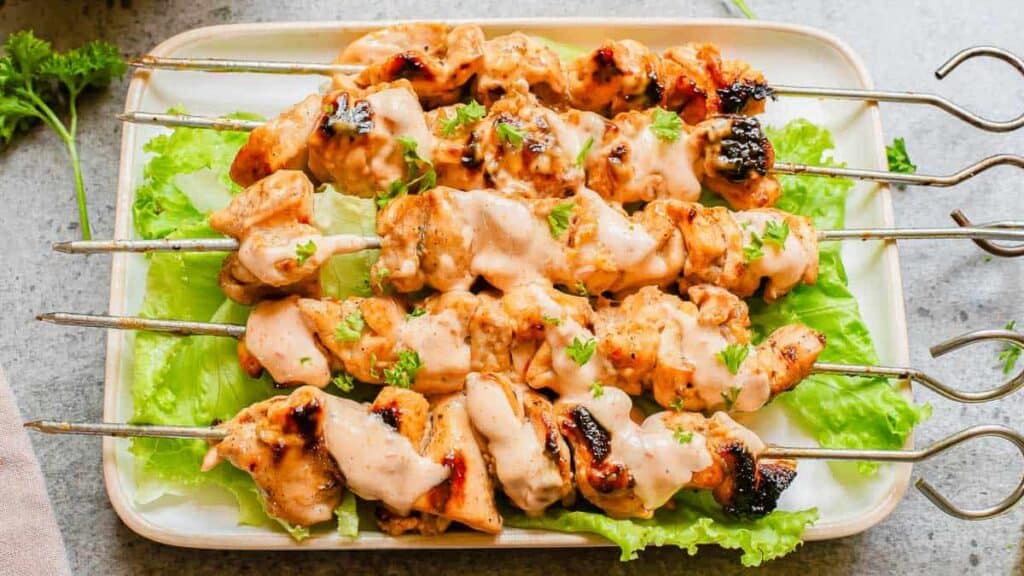 Grilled chicken skewers with sauce are served on a bed of lettuce on a rectangular plate.