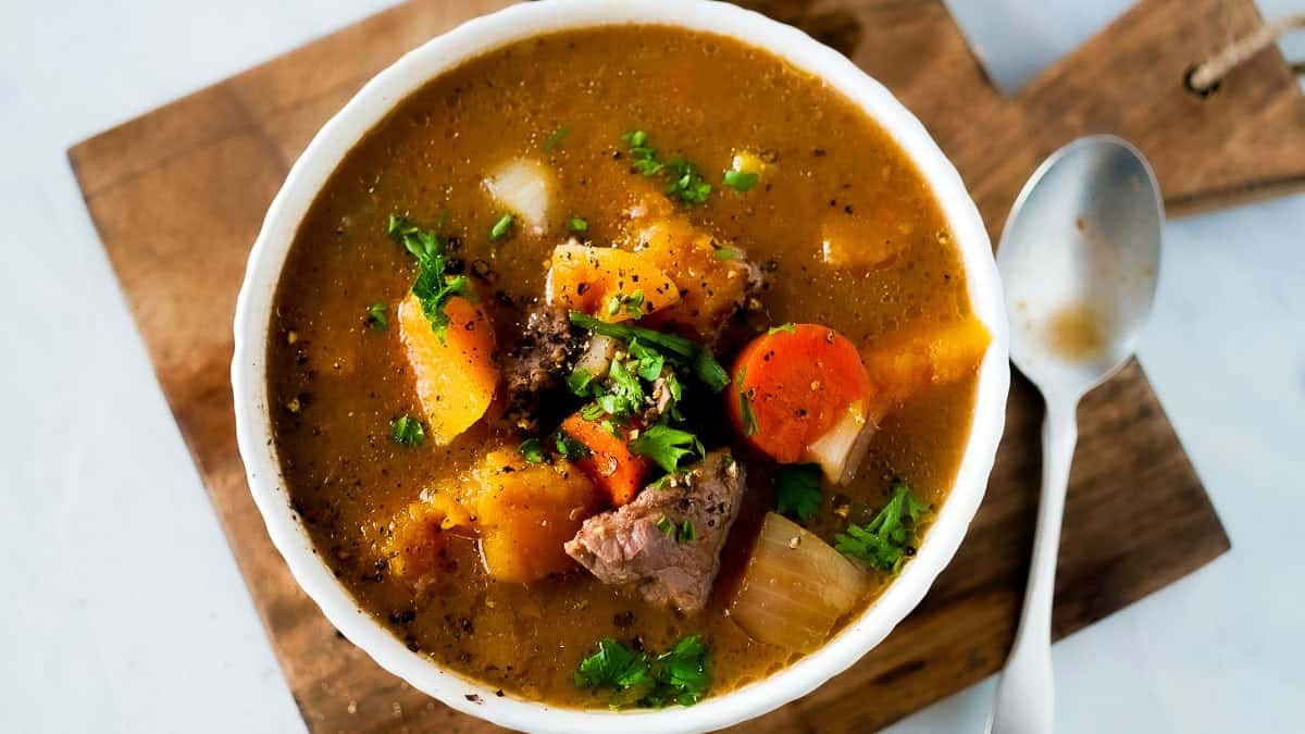A bowl of chunky beef stew with carrots, onions, and herbs, served on a wooden board.