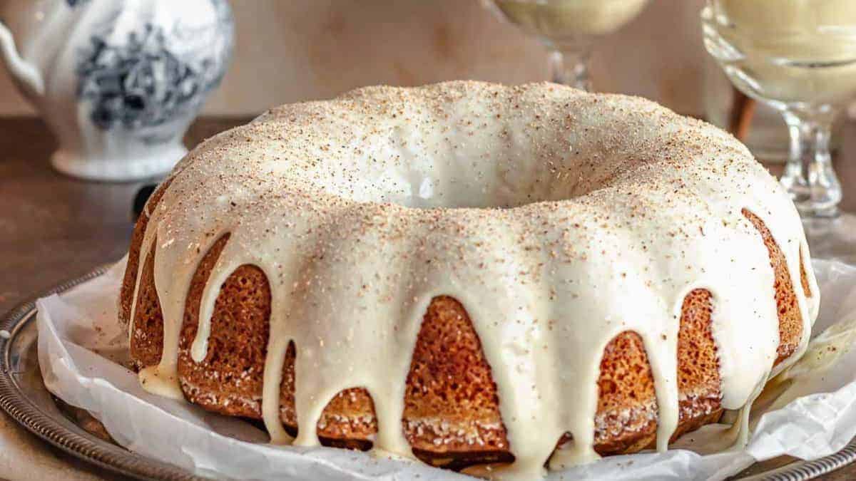 A Bundt cake with white glaze drips sits on a silver tray. Tea cups and a teapot are in the background.
