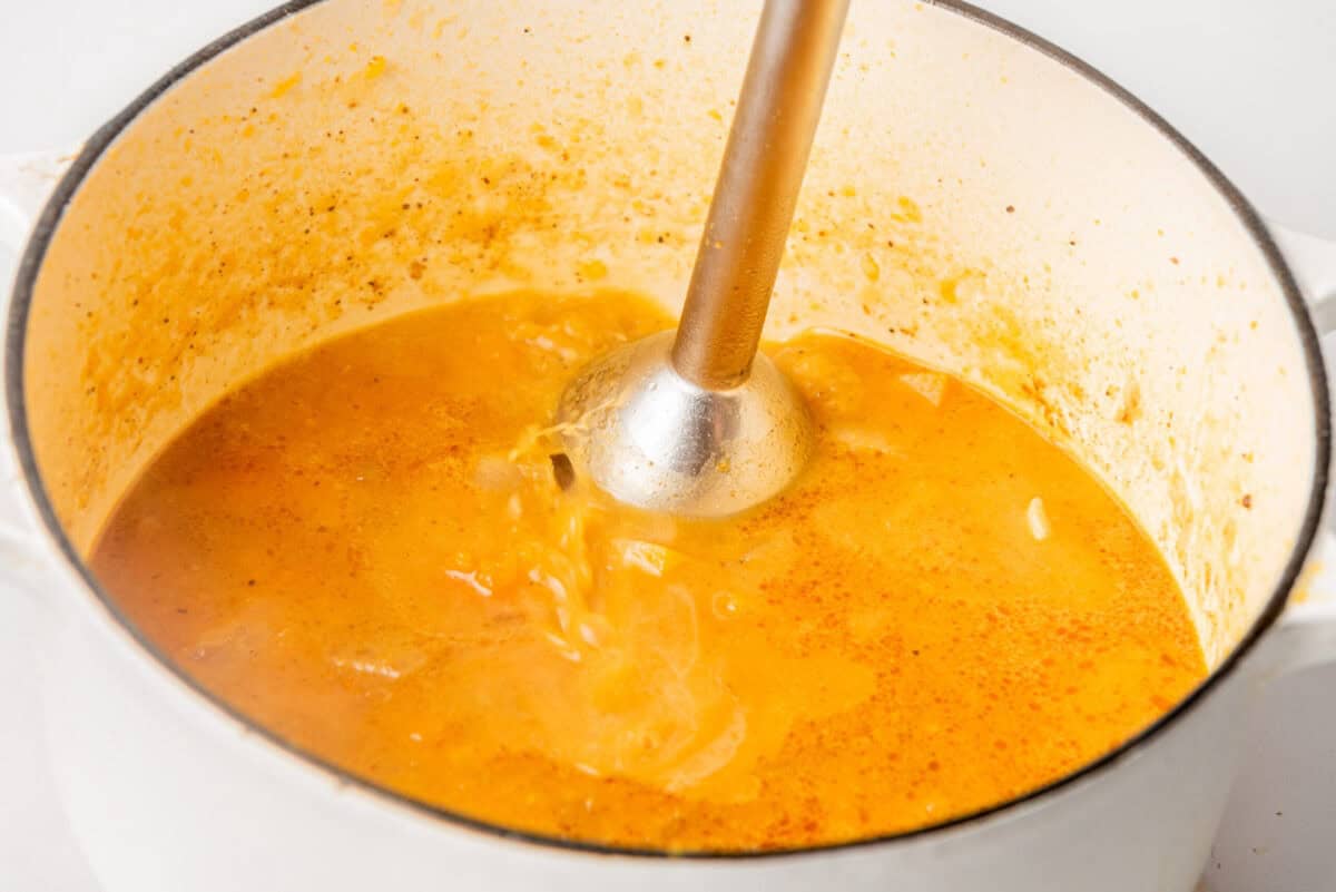 An immersion blender mixing orange soup in a white pot.