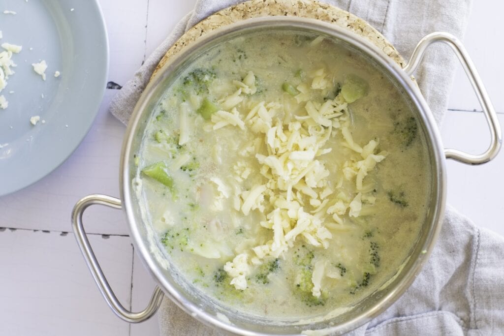 A pot of creamy broccoli soup topped with shredded cheese, placed on a cork mat.