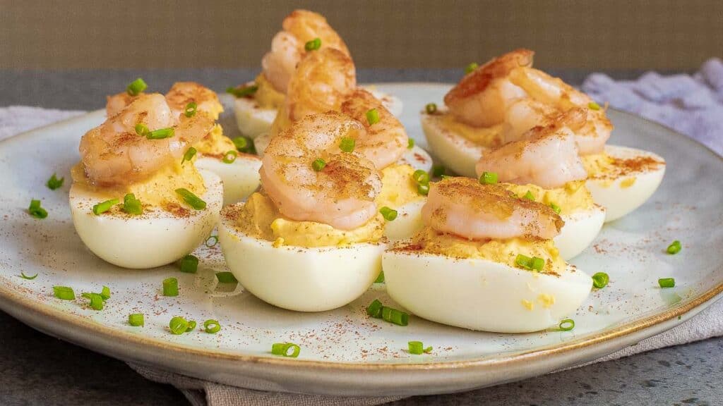 Plate of deviled eggs topped with cooked shrimp and sprinkled with chopped chives and paprika.