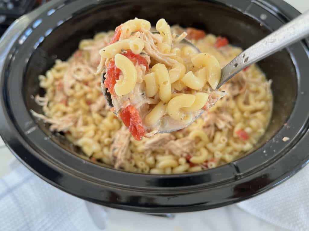 Close-up of a spoonful of macaroni with shredded chicken and tomatoes being lifted from a slow cooker filled with the same mixture.