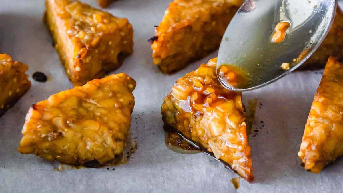 Close-up of triangular tempeh pieces on parchment paper, being glazed with sauce from a spoon.