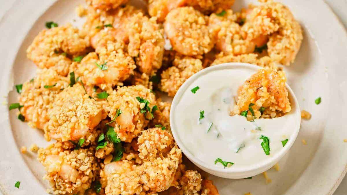 A plate of breaded popcorn shrimp garnished with chopped herbs, accompanied by a bowl of creamy dipping sauce.