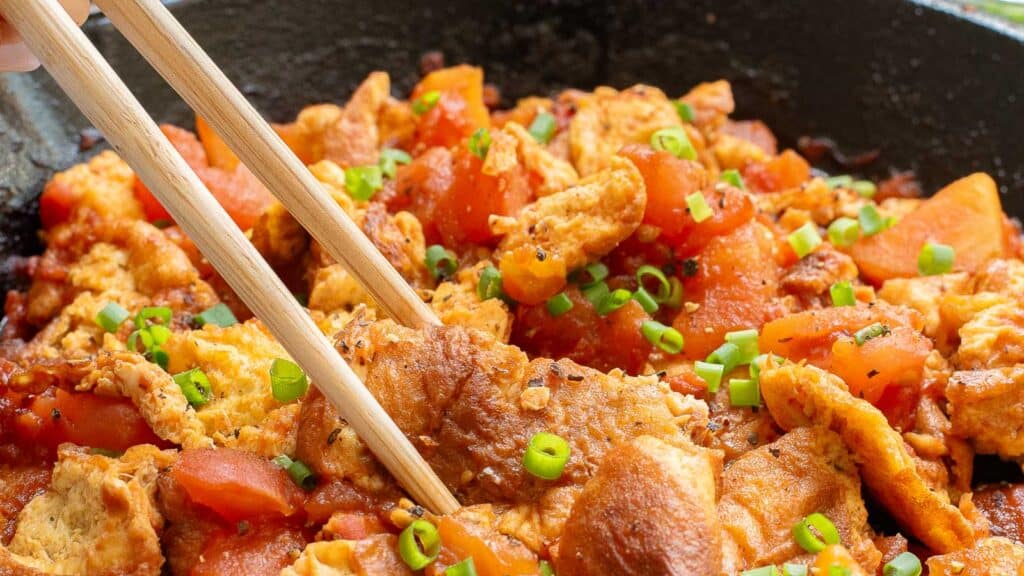 Chopsticks picking up stir-fried tofu with tomatoes and green onions in a skillet.