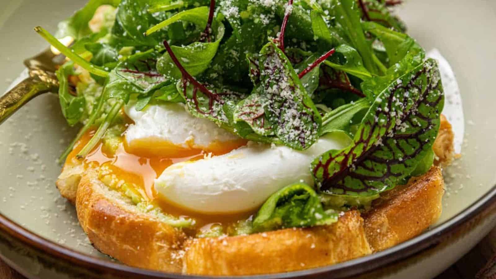 A poached egg on toast with greens, accompanied by creamy avocado and topped with grated cheese, served on a plate.