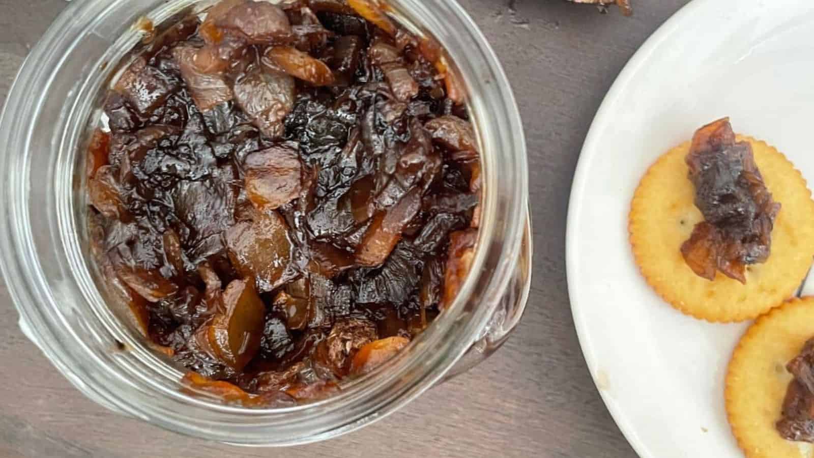 A jar of dark balsamic onion relish sits next to a plate with crackers generously topped with the same rich jam, all elegantly placed on a rustic wooden surface.