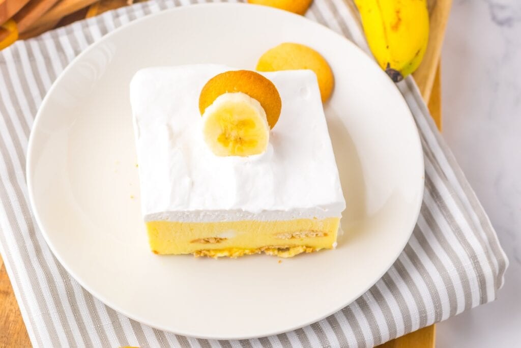 A slice of banana pudding topped with whipped cream, a banana slice, and a cookie on a white plate with a striped napkin.