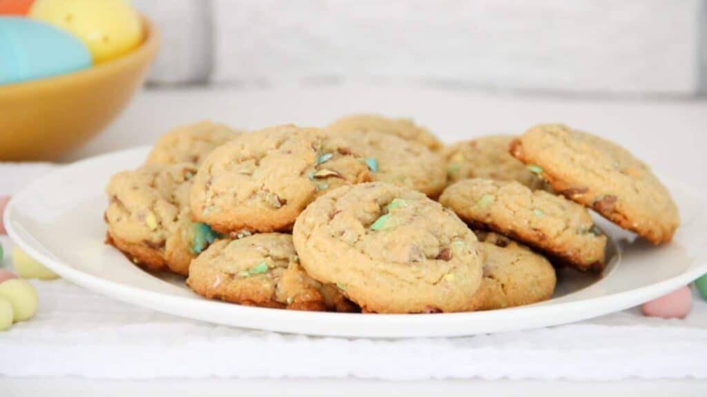 A white plate with a stack of chocolate chip cookies with colorful candy pieces. A bowl with colorful eggs is in the background.