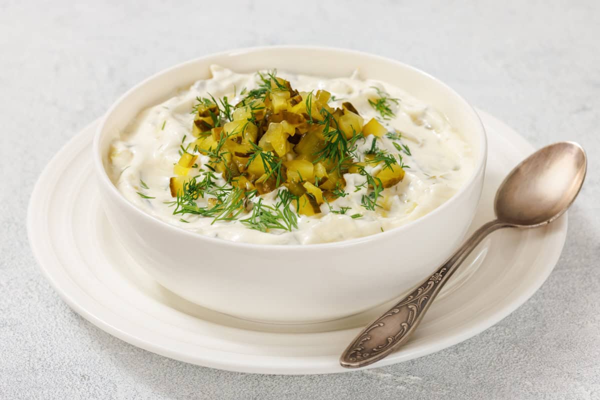 A bowl of Dill Pickle Dip with chopped pickles and dill on top, placed on a white plate. A spoon rests beside the bowl.