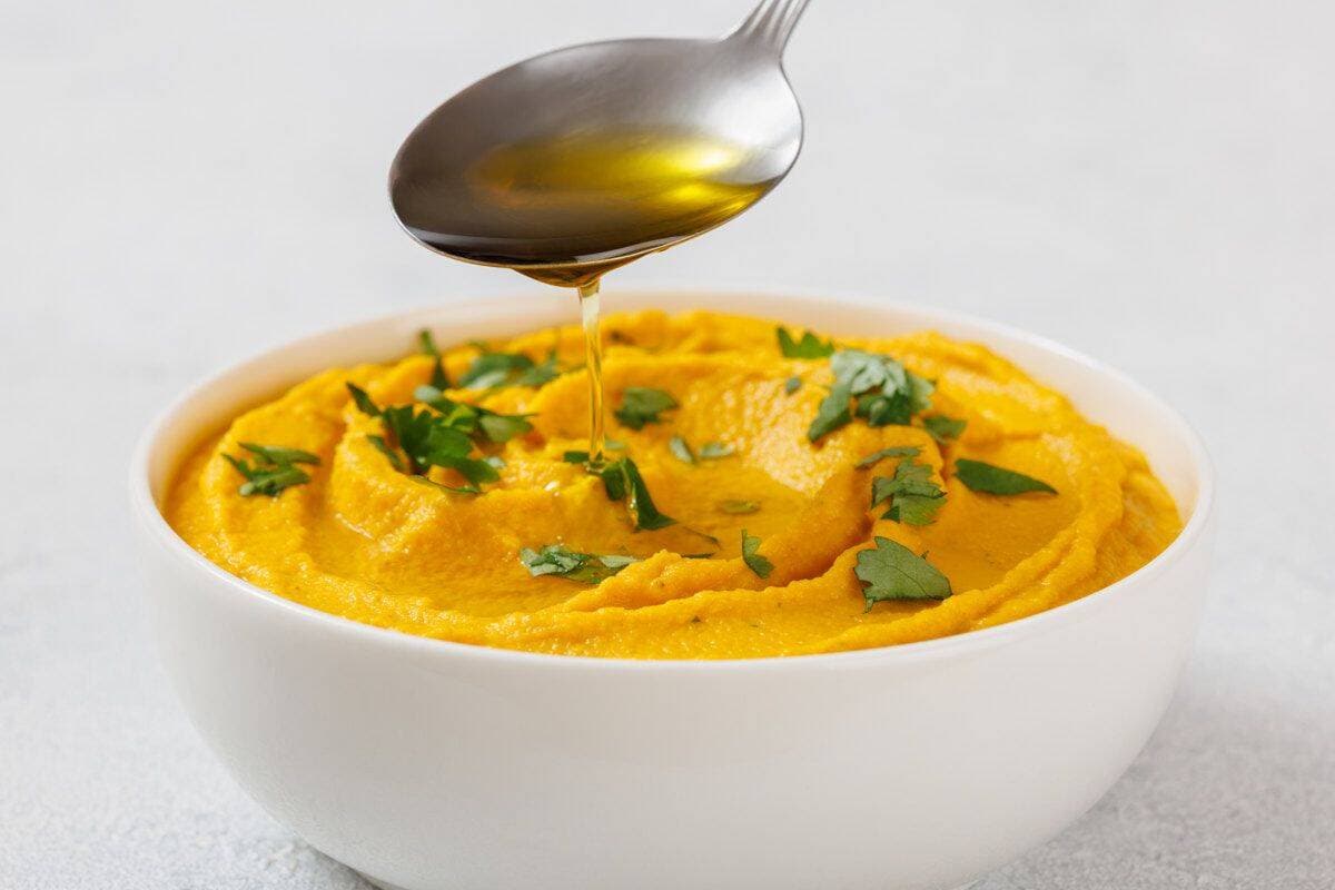 Bowl of Roasted Carrot Hummus garnished with green herbs, with a spoon drizzling oil above it.