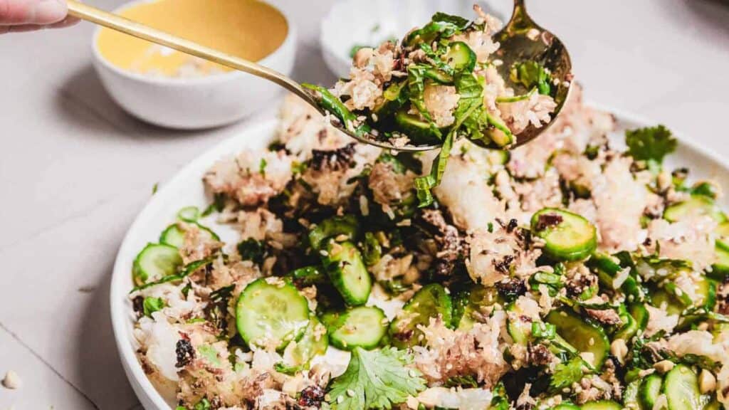 Close-up of a hand serving a mixed salad with bold flavors, featuring fresh ingredients like cucumbers, herbs, nuts, and grains on a white plate. A small yellow bowl is in the background.