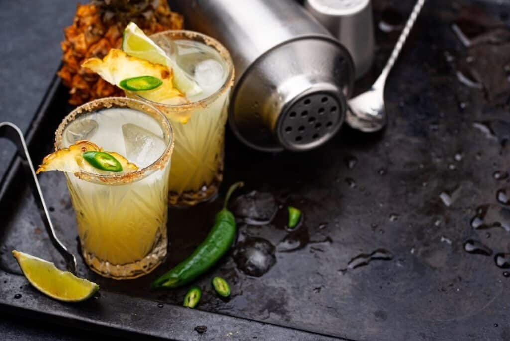 Two iced cocktails with pineapple slices and chili garnishes sit on a dark tray. Beside them are a cocktail shaker, lime wedge, and green chilies.