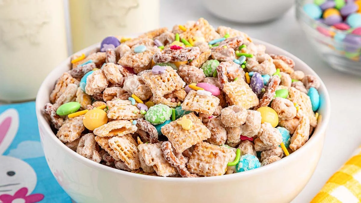 A bowl of colorful snack mix containing Chex cereal, chocolate candies, and pretzels sits on a table. Two bottles of milk and another bowl of candies are visible in the background.