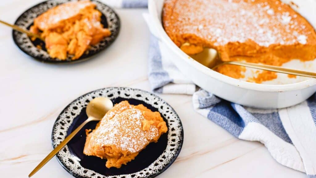 Two servings of sweet potato casserole dusted with powdered sugar on plates, with a serving dish and a blue-and-white towel nearby. Two spoons are placed on the plates.