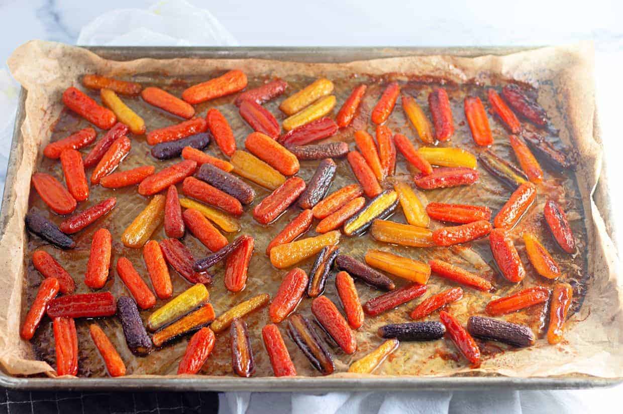 A baking sheet lined with parchment paper holds honey-roasted baby carrots in vibrant orange, yellow, and purple hues. The carrots are glistening with a caramelized glaze.