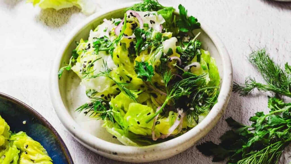 A bowl of green salad featuring bold flavors and fresh ingredients like lettuce, parsley, dill, red onion, and black sesame seeds sits on a light background.