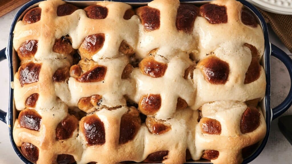 A baking dish filled with freshly baked hot cross buns featuring a shiny glaze and visible raisins.