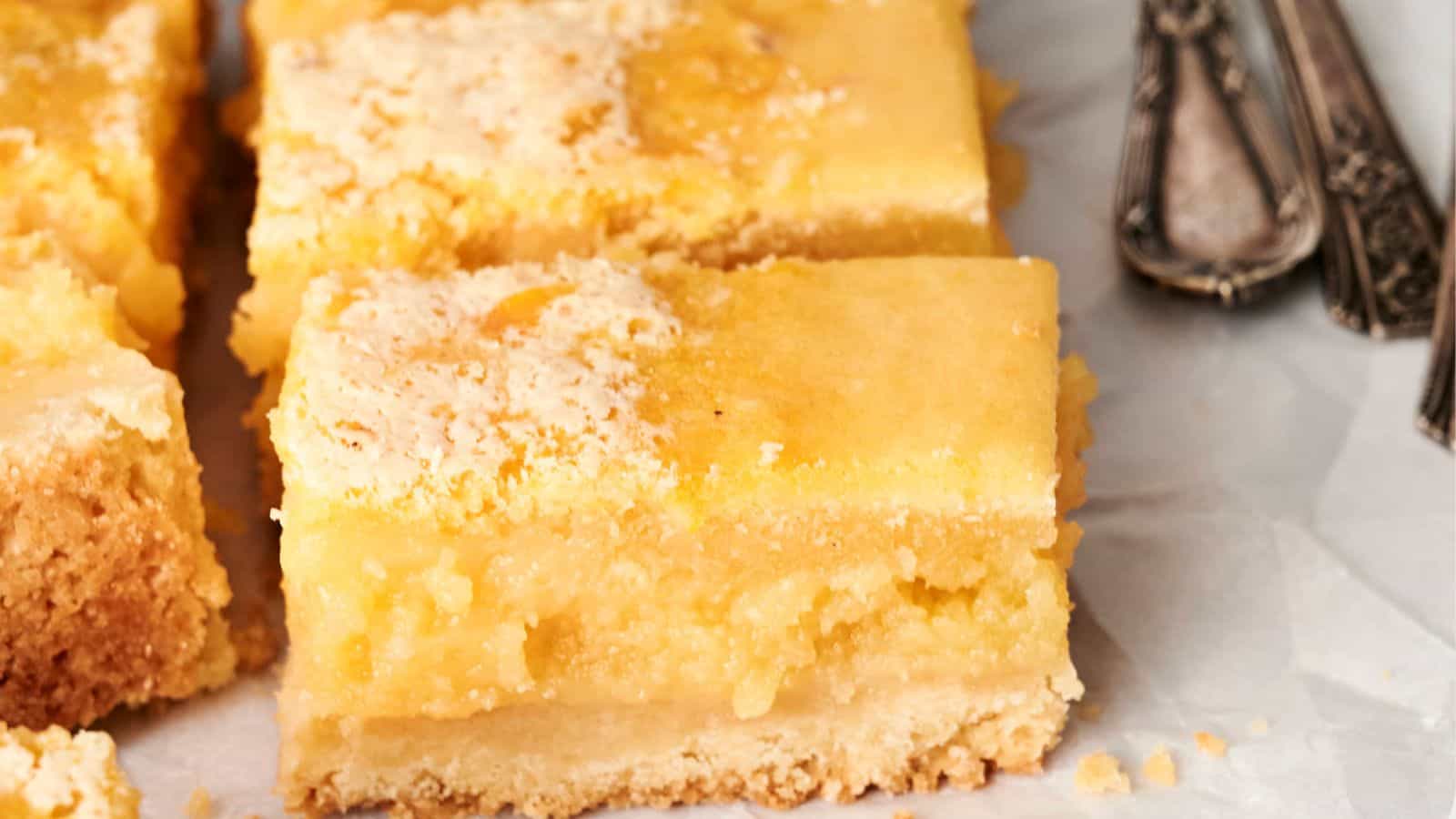 Close-up of lemon bars with a crumbly base and a smooth, yellow filling, cut into squares, placed on parchment paper. Silver utensils are visible to the side.