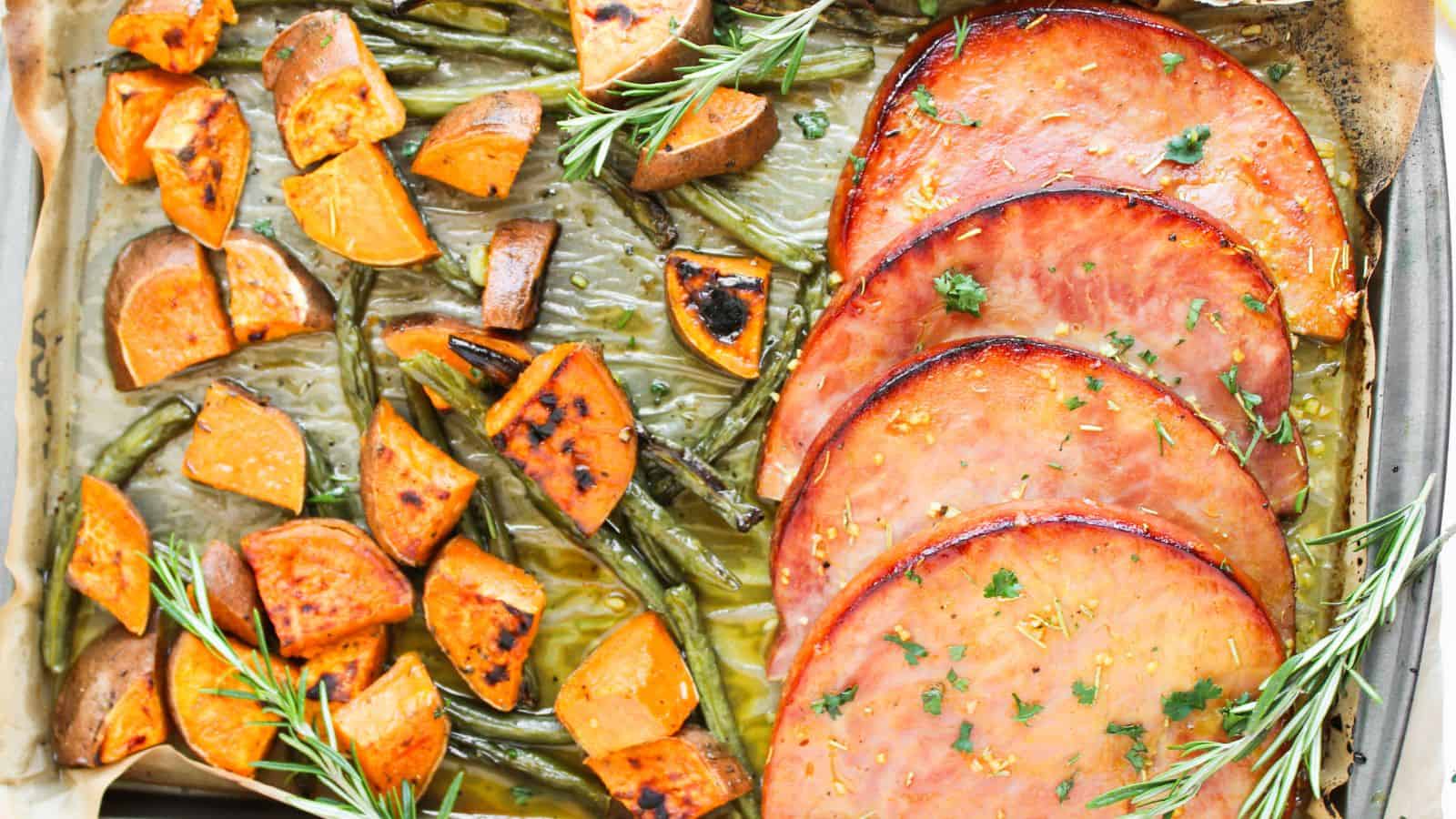 A baked dish with slices of glazed ham, roasted sweet potato cubes, and green beans, garnished with rosemary sprigs.