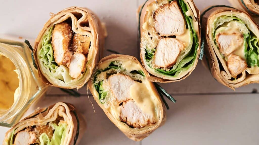 Close-up of sliced wraps filled with grilled chicken, lettuce, and sauce, arranged on a light surface.