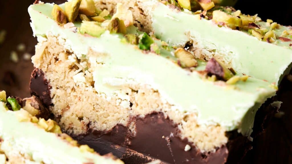 Close-up of slices of a dessert with layers of green frosting, crumbly base, chopped pistachios on top, and a thick dark chocolate layer at the bottom.
