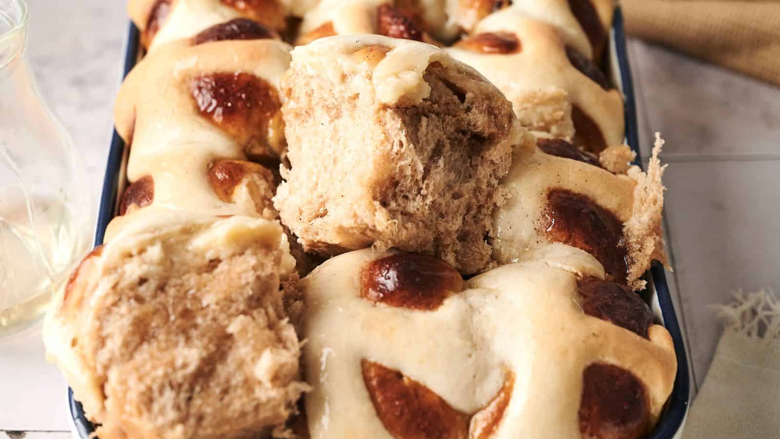 A tray of freshly baked hot cross buns with golden brown tops and a soft, fluffy texture. One bun is partially broken to show its interior.