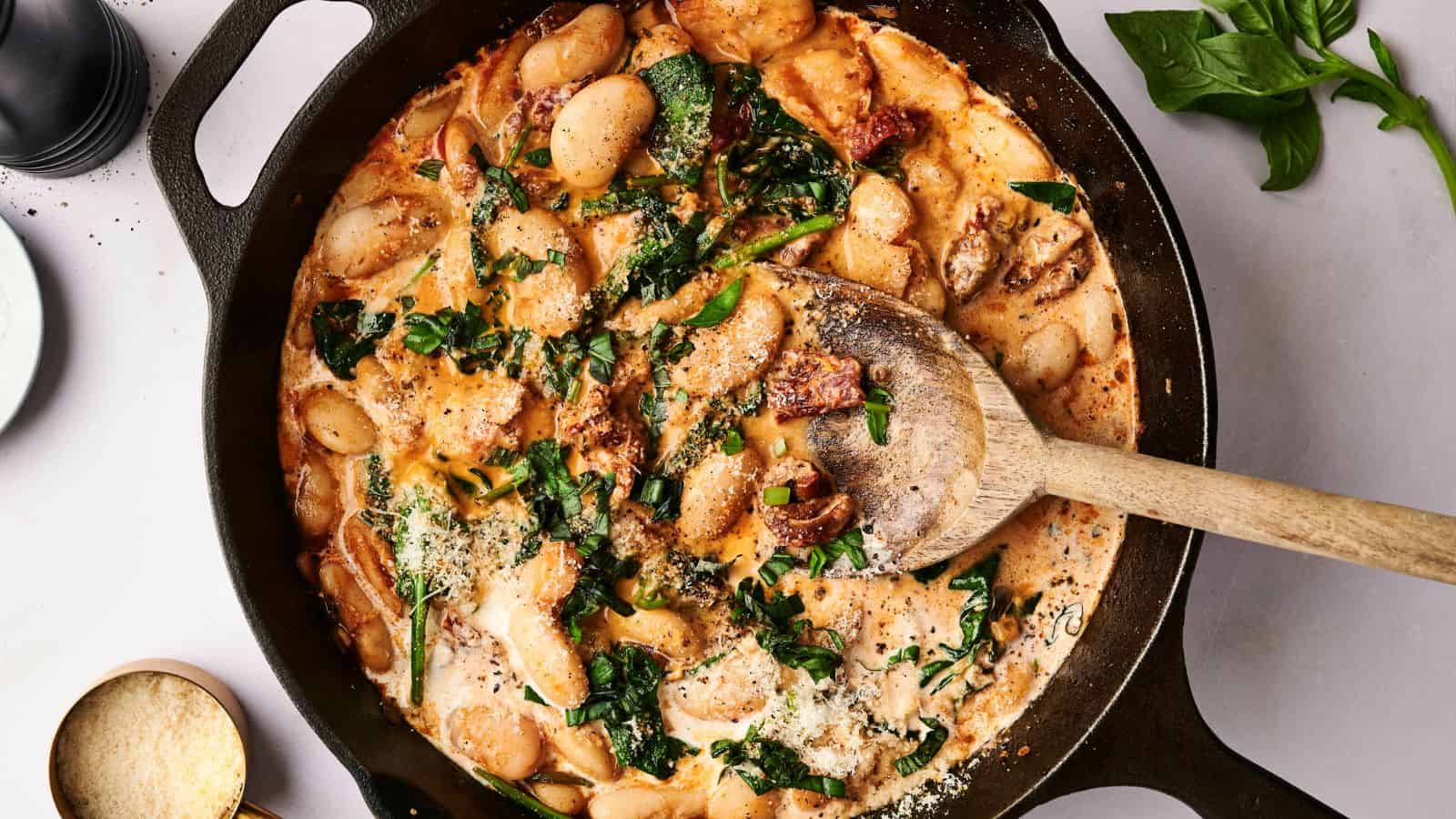 A cast iron skillet with creamy pasta mixed with spinach and sun-dried tomatoes, topped with cheese, with a wooden spoon resting inside.