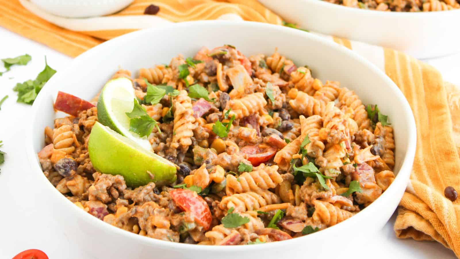 A bowl of fusilli pasta mixed with ground beef, black beans, chopped tomatoes, and herbs, garnished with lime wedges.