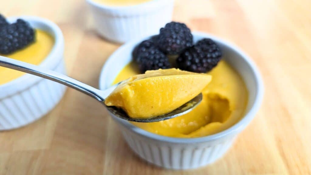 A spoon holds a portion of mango mousse above a ramekin, topped with blackberries. Other ramekins filled with the yellow dessert are visible in the background.