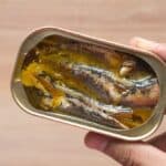 A hand holding an open tin of sardines in oil against a wooden background.